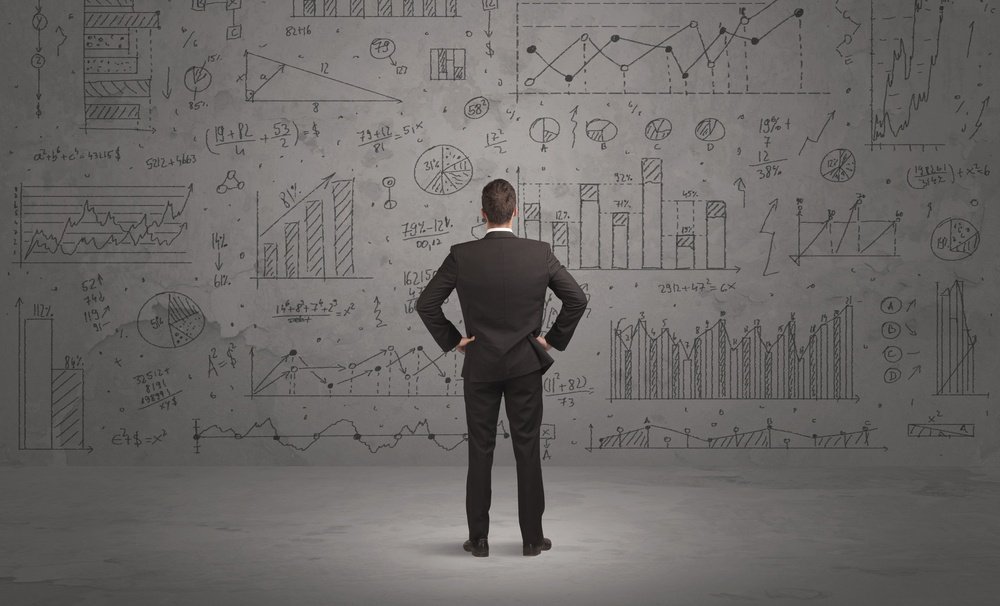 A successful confident businessman thinking about decisions, standing in front of wall full with graph pie charts and calculations concept.jpeg